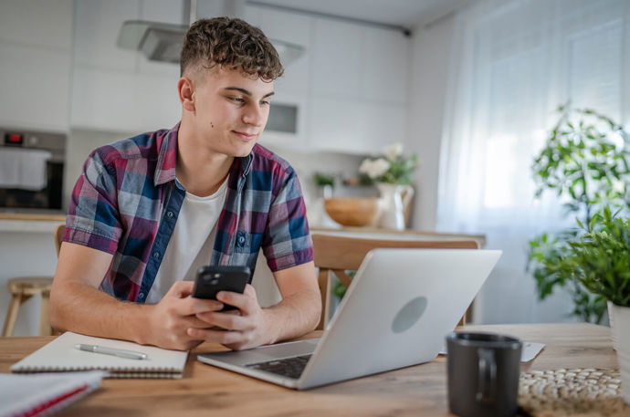 A young man looking on his mobile while working on a laptop