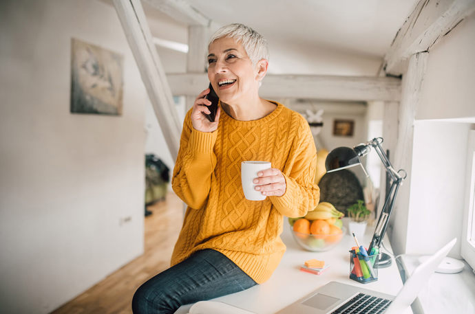 A smiling woman sitting on a desk, holding a hot drink while speaking on the phone